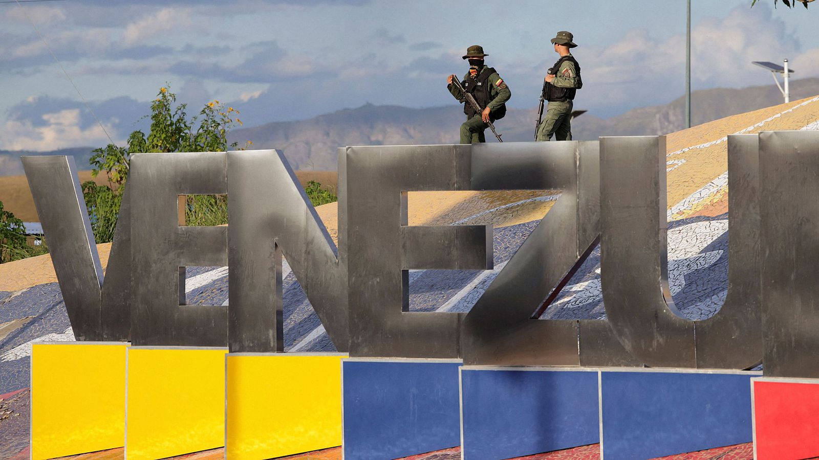 Dos guardias nacionales venezolanos, vestidos con uniformes militares y portando armas, se encuentran en la frontera, sobre una estructura con la palabra "VENEZUELA" escrita en grandes letras y con los colores de la bandera.