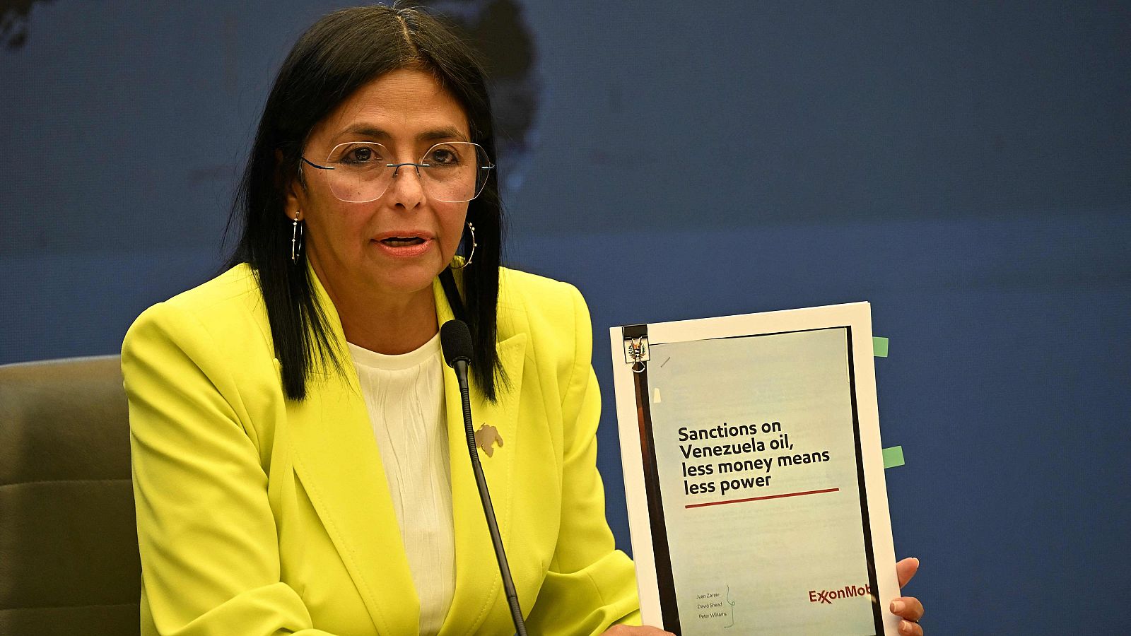 Delcy Rodríguez, vicepresidenta de Venezuela, aparece en una conferencia de prensa, sosteniendo un documento con el logo de una empresa petrolera y notas adhesivas, vestida con un blazer amarillo.