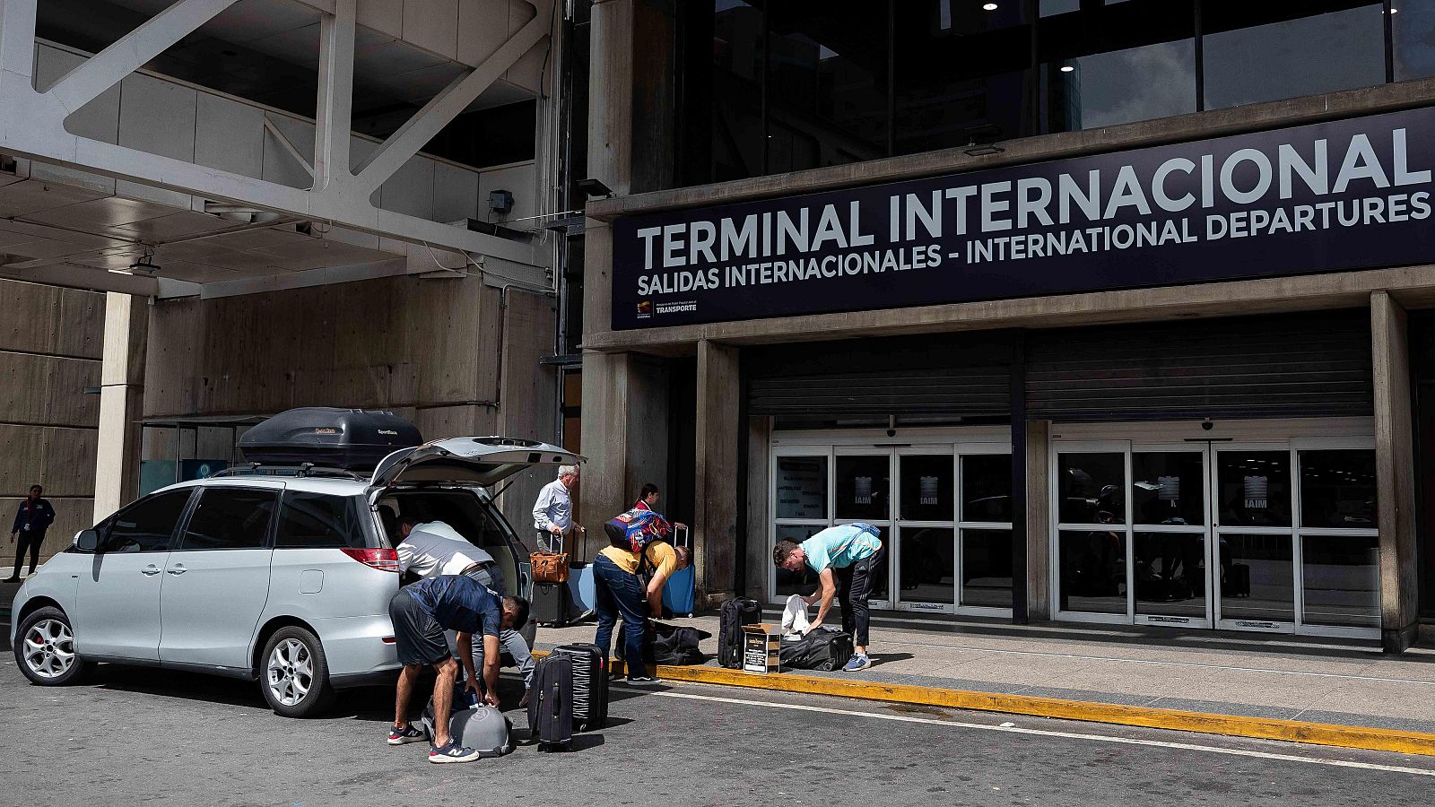 En la entrada de una terminal internacional, un monovolumen gris es el foco de atención mientras personas descargan equipaje. Maletas en el suelo y un letrero que indica la dirección del área.