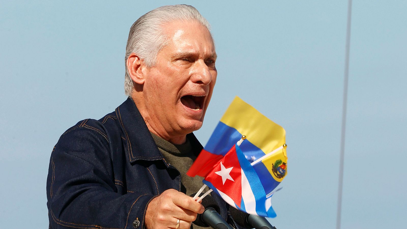 Miguel Díaz-Canel, con chaqueta vaquera, habla frente a un micrófono sosteniendo banderas. Se encuentra en un acto público con un fondo celeste claro.