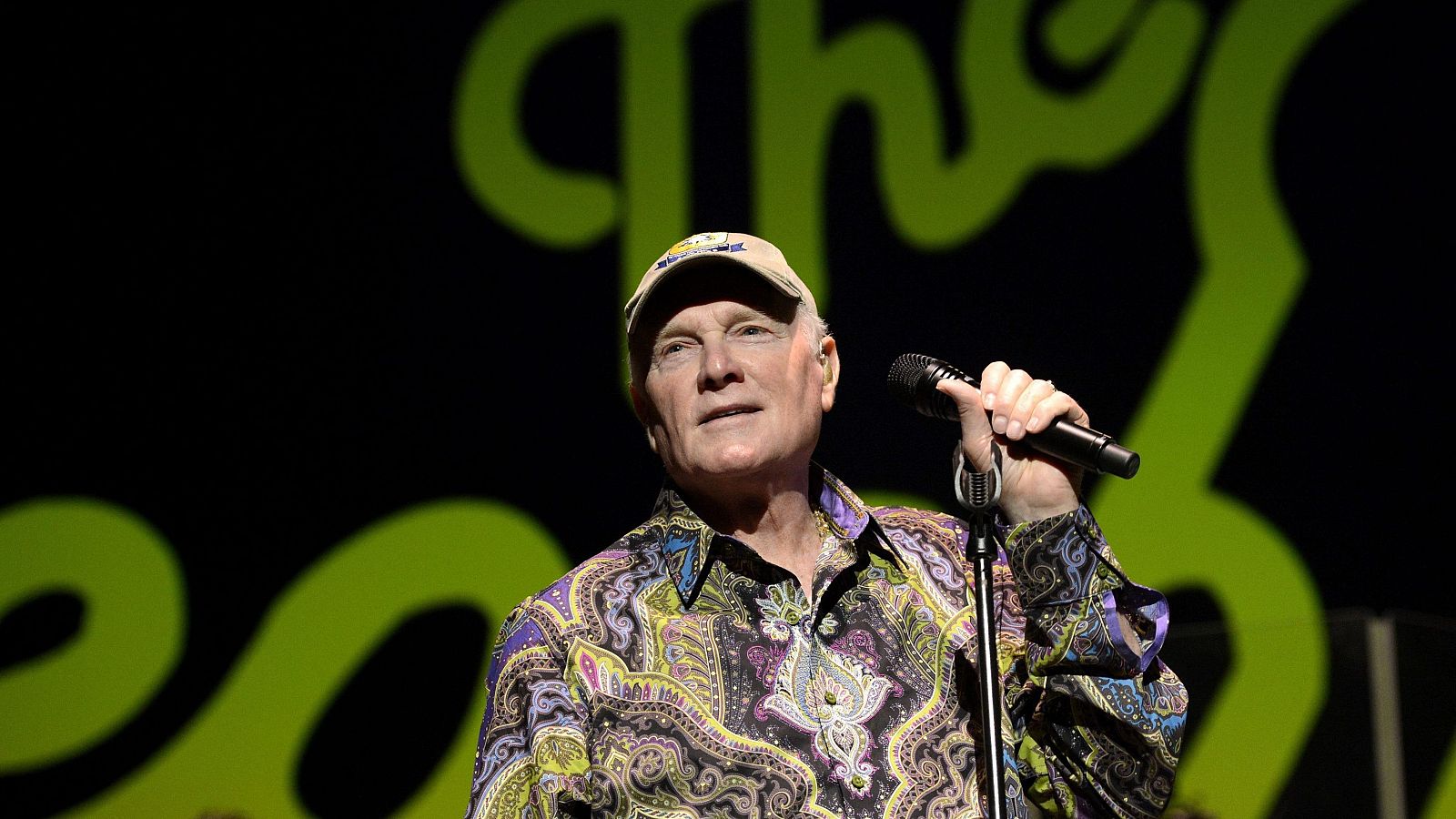 En un escenario oscuro, Mike Love, con una gorra y camisa estampada, canta con un micrófono en la mano. El logo de la banda, en verde lima, se vislumbra en el fondo.