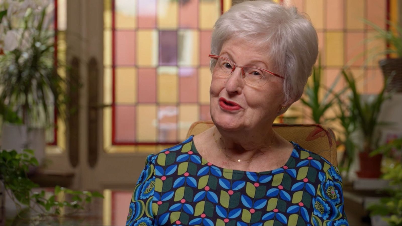 Retrato de una mujer mayor con gafas rojas y labios pintados, vestida con una blusa floreada. El fondo presenta una ventana con vidrieras de colores.