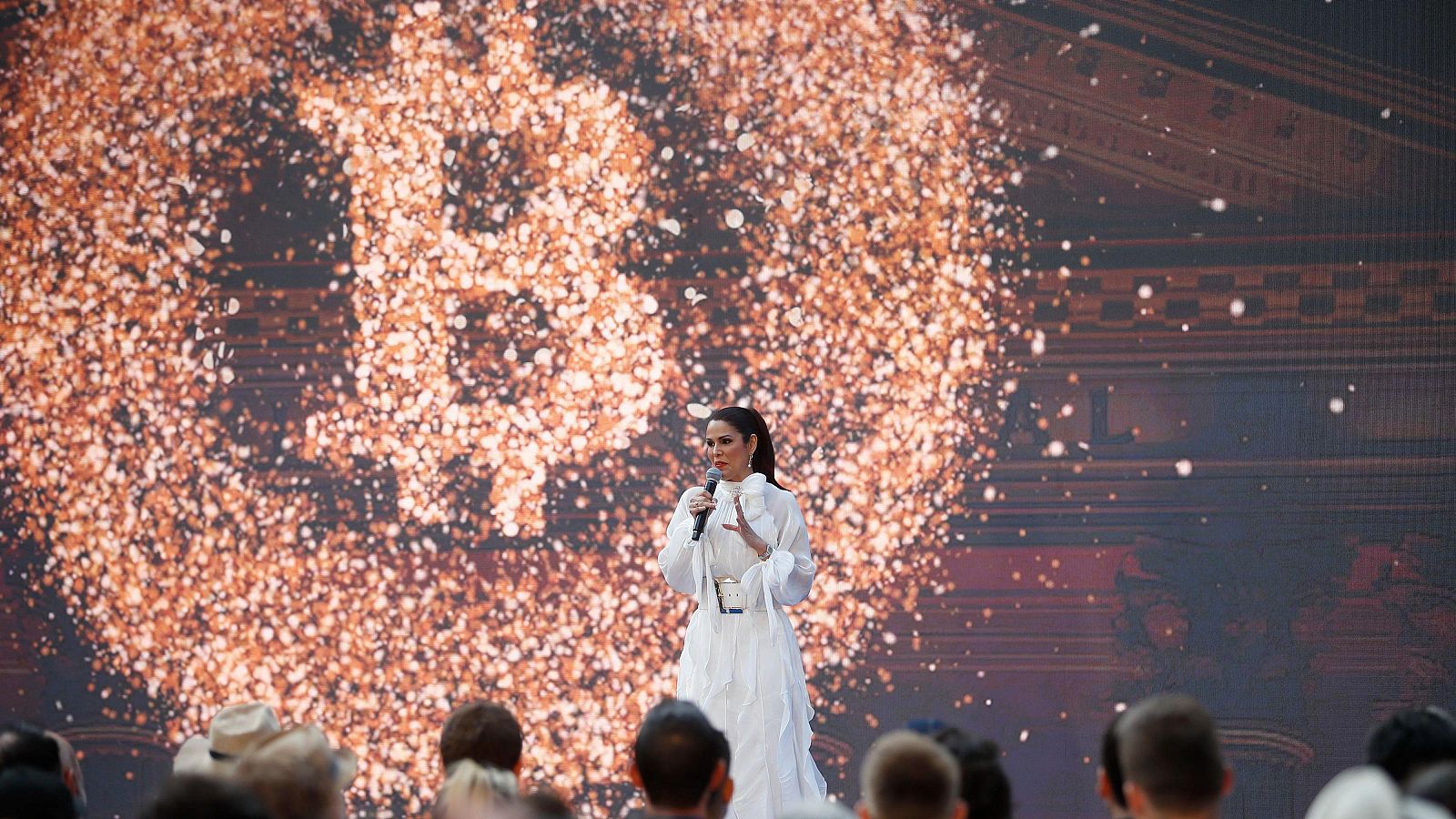La embajadora, vestida de blanco, habla ante un micrófono con un logo de Bitcoin proyectado detrás, en un evento con audiencia presente.