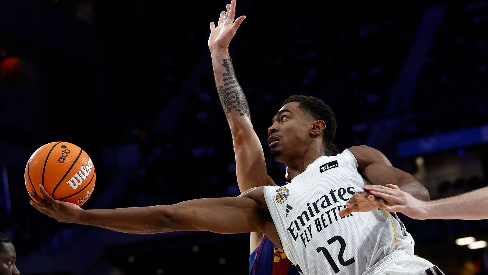 En la imagen, Théo Maledon, con la camiseta del Real Madrid y el número 12, se dispone a lanzar el balón de baloncesto, sujetando un balón naranja.