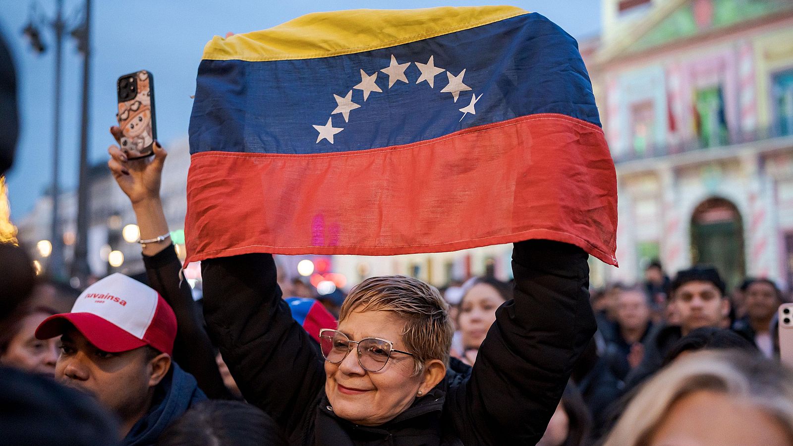 En una concentración en Madrid, una mujer sonríe mientras sostiene la bandera de Venezuela, con sus colores característicos y ocho estrellas, frente a una multitud de personas.