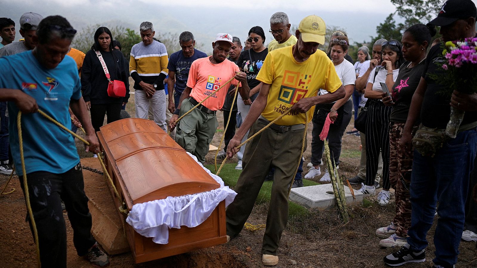 En un cementerio, un grupo de personas asiste al entierro de un ser querido. El ataúd de madera es descendido a la tumba, mientras algunos presentes expresan su dolor y una persona sostiene un ramo de flores.