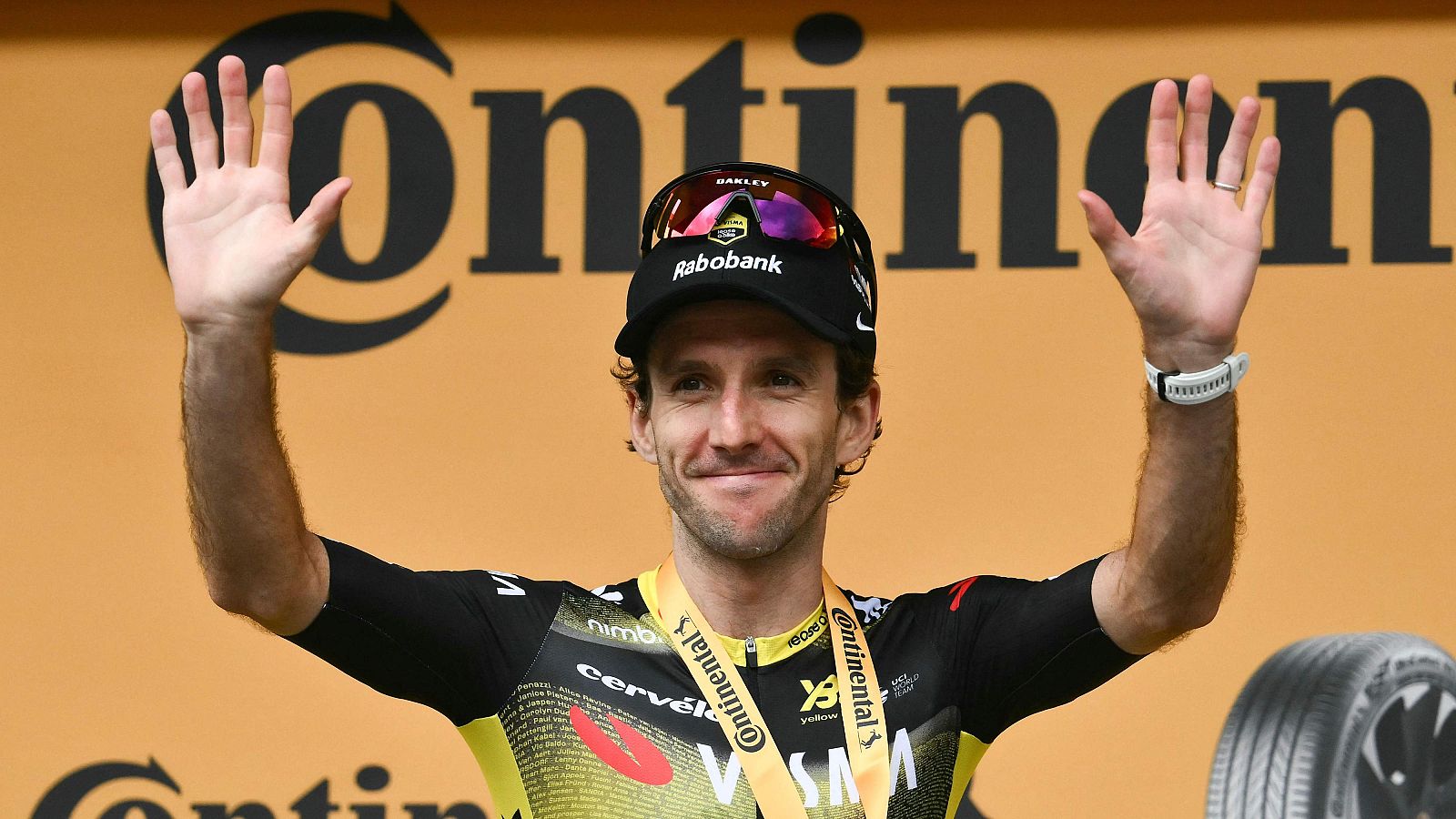Un ciclista, posiblemente Simon Yates, celebra una victoria con los brazos en alto, vistiendo un maillot con logotipos y una gorra. Lleva gafas de sol y una medalla, con un reloj deportivo en la muñeca.