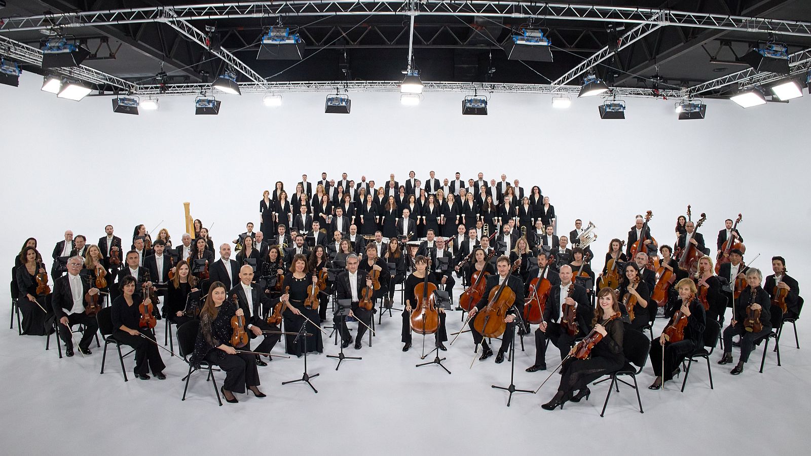 En un estudio con fondo blanco, la Orquesta y Coro de RTVE, vestidos de negro, interpretan música. El coro se ubica atrás y la orquesta delante, con instrumentos como violines y trompetas.