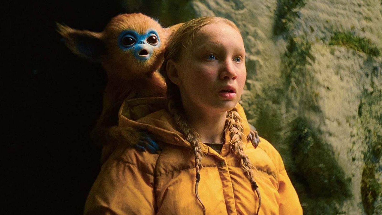 Una joven rubia con trenzas y chaqueta amarilla, con expresión de asombro, comparte el encuadre con una criatura fantástica de pelaje marrón y rasgos azules, en un entorno rocoso y natural.
