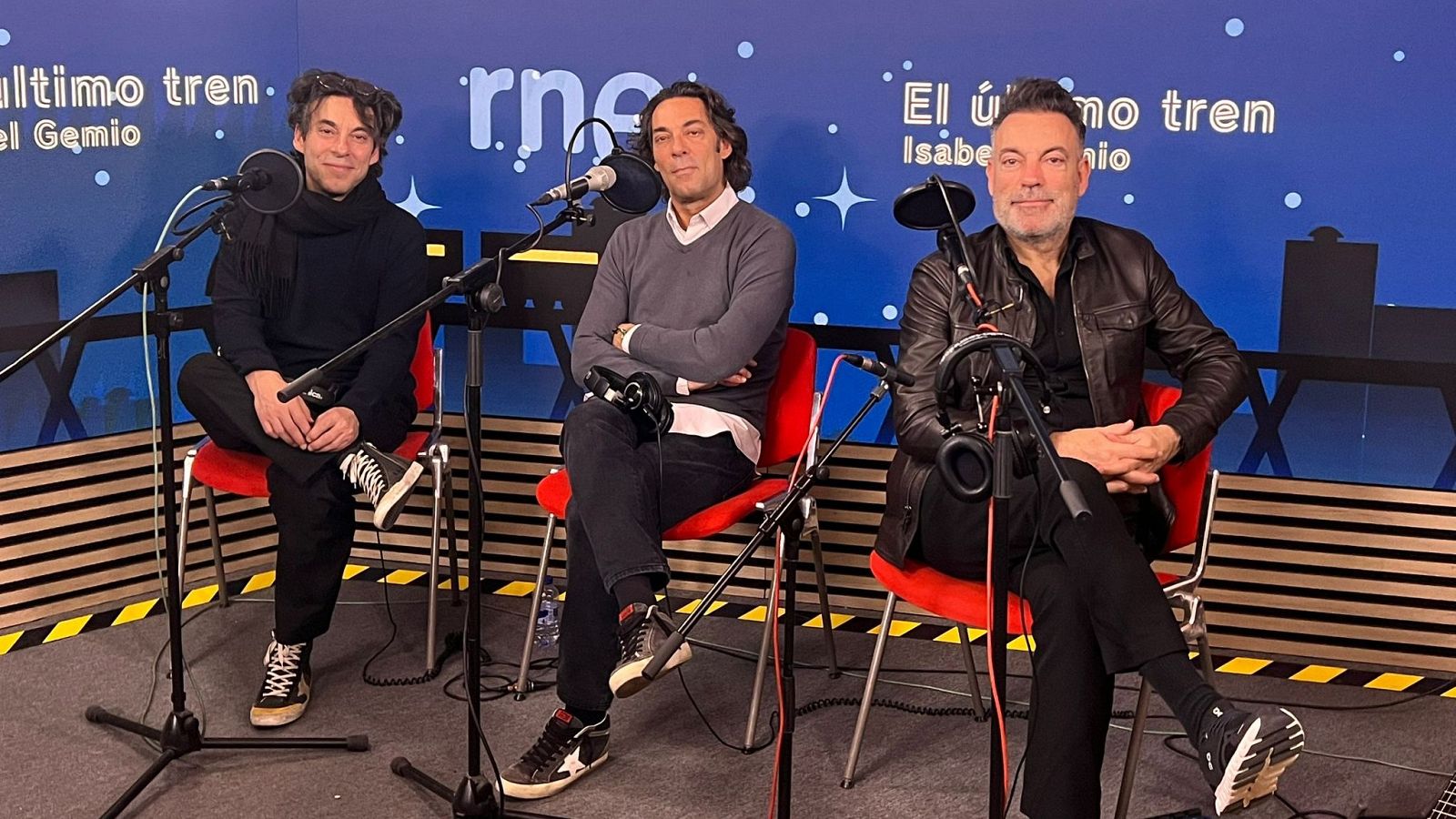 Los tres integrantes de Café Quijano, vestidos con prendas oscuras, se encuentran en un estudio de radio con micrófonos, posiblemente para una entrevista o grabación. Se observa el logo de una emisora y el título de un programa.