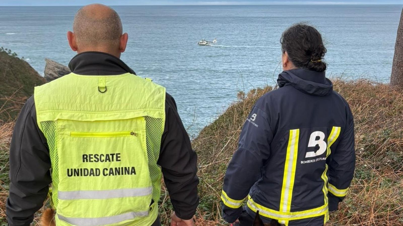En un acantilado, dos personas de espaldas, posiblemente de un equipo de rescate, observan el mar. Uno lleva un chaleco con la inscripción "RESCATE UNIDAD CANINA" y el otro una chaqueta con el logo de "B BOMBEROS".