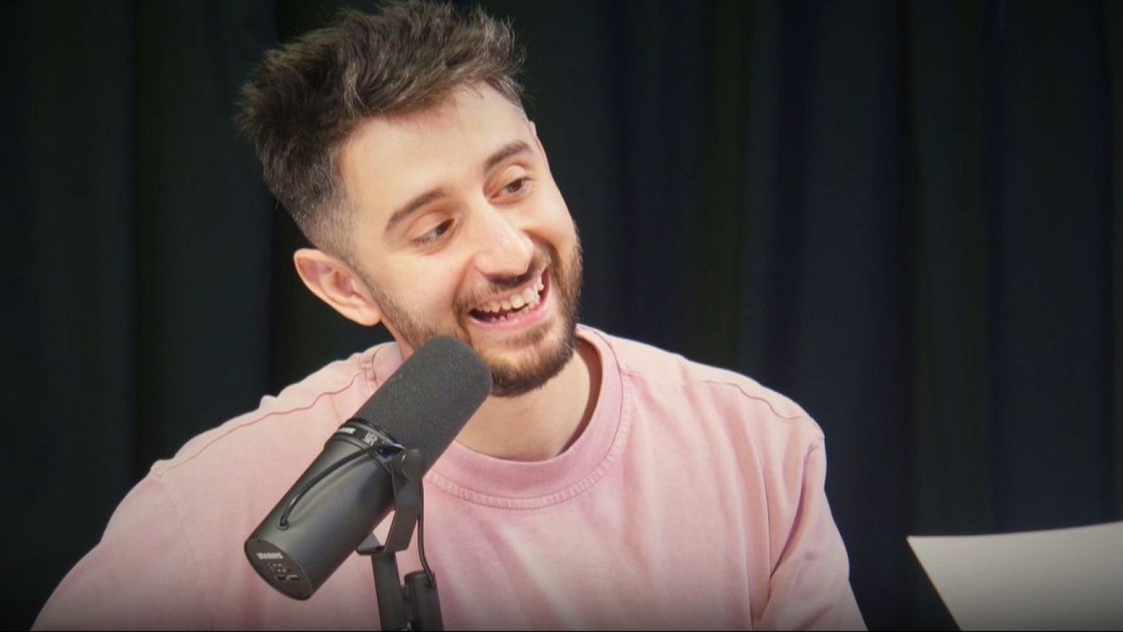 Un hombre con barba y camiseta rosa sonríe frente a un micrófono, posiblemente en un estudio de radio. El fondo es una cortina oscura, y se observa el texto "yorya 2" y "Radio".