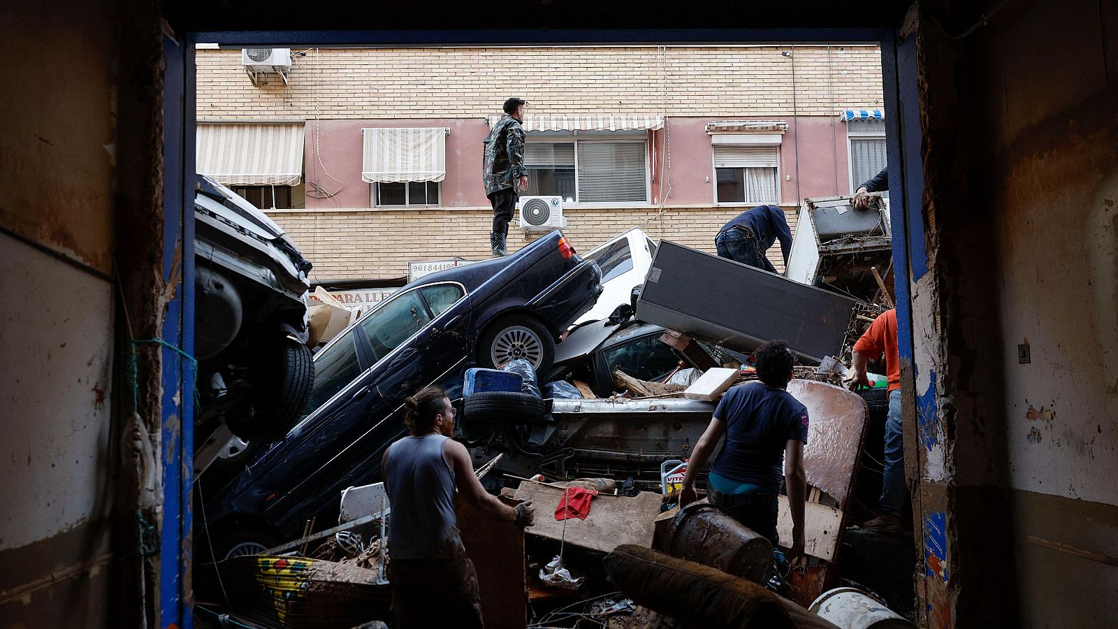 Una calle urbana se muestra completamente inundada y llena de escombros, con coches amontonados y personas caminando entre los restos, evidenciando una situación de caos y destrucción.