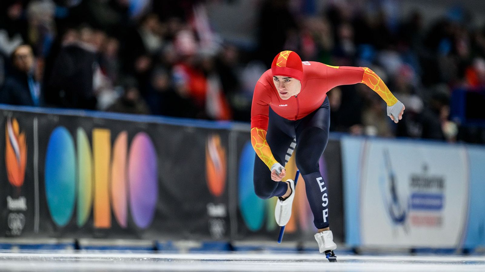 Nil Llop, patinador de velocidad, compite en una prueba de 500 metros, vistiendo un traje rojo con detalles en amarillo y negro, y la inscripción "ESP". Se desliza sobre la pista de hielo con la mirada fija en la meta, con público al fondo.