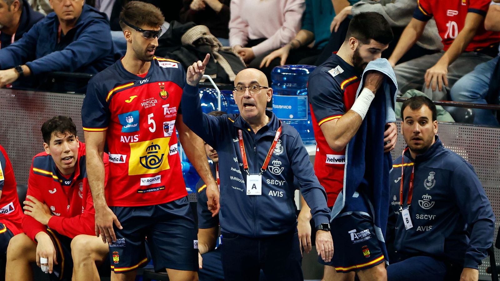 Jordi Ribera, entrenador de balonmano, da indicaciones a sus jugadores, quienes visten uniformes deportivos. Se observan detalles como gafas, chándal con logos y botellas de agua, sugiriendo un ambiente de competición.
