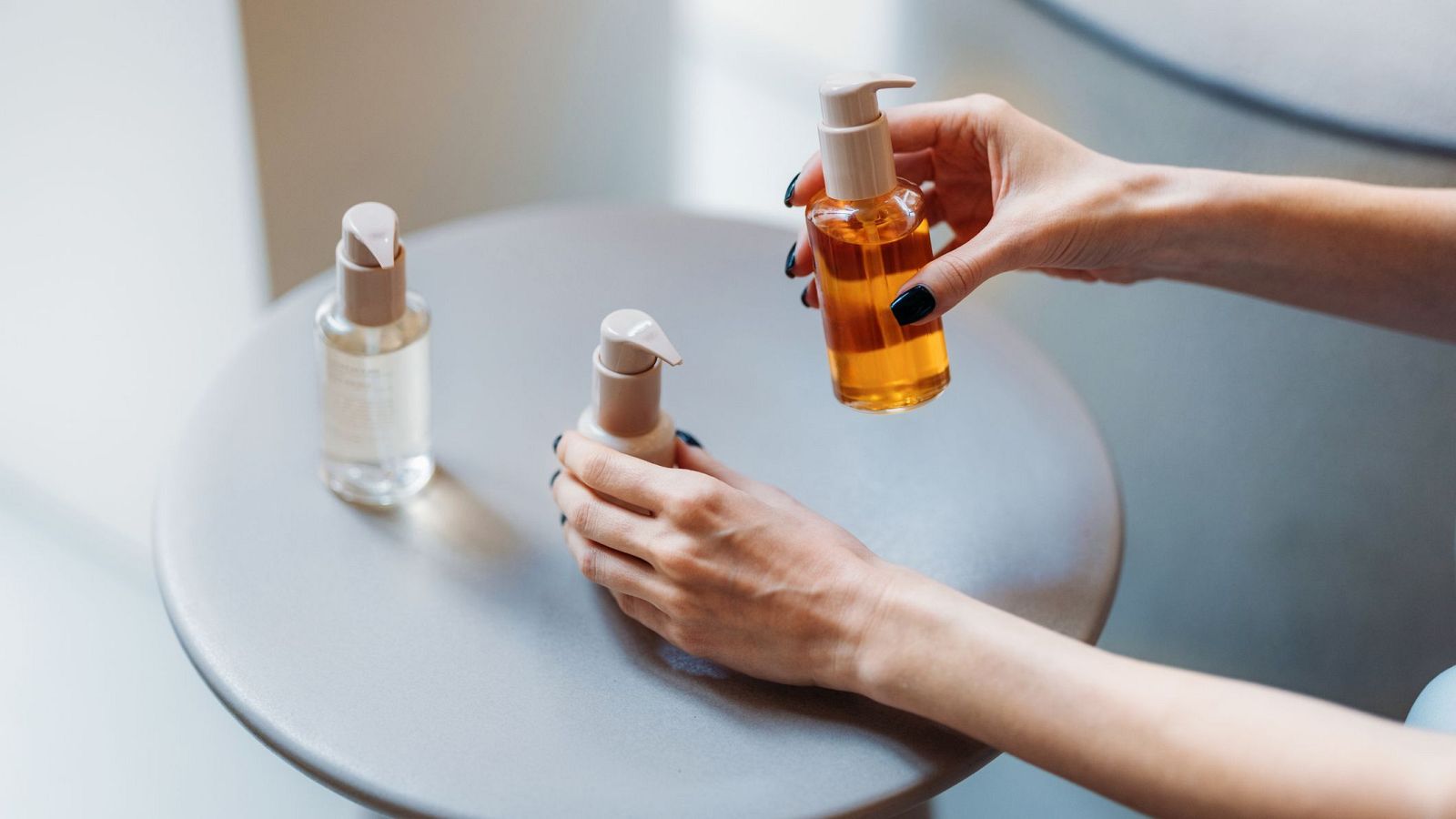 Tres botellas de cosméticos, con dispensadores y líquidos de distintos colores, se exhiben sobre una mesa. Una mano con uñas oscuras sostiene una de las botellas en el aire, mientras que las otras dos reposan sobre la mesa.