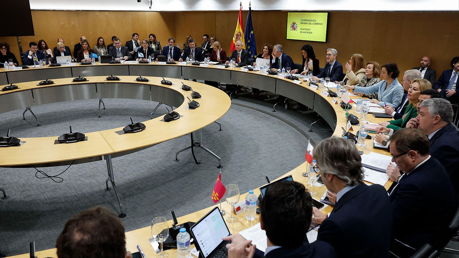 Una reunión formal con múltiples personas sentadas alrededor de una mesa redonda equipada con micrófonos individuales. Se pueden observar banderas y un monitor con texto.