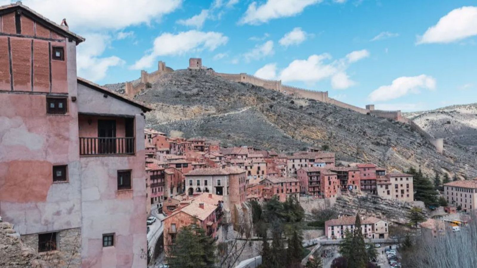 Una panorámica de Teruel, España, muestra edificios de arquitectura tradicional en tonos rojizos y ocres, con balcones de madera. Una muralla medieval con torres domina el paisaje, bajo un cielo azul.