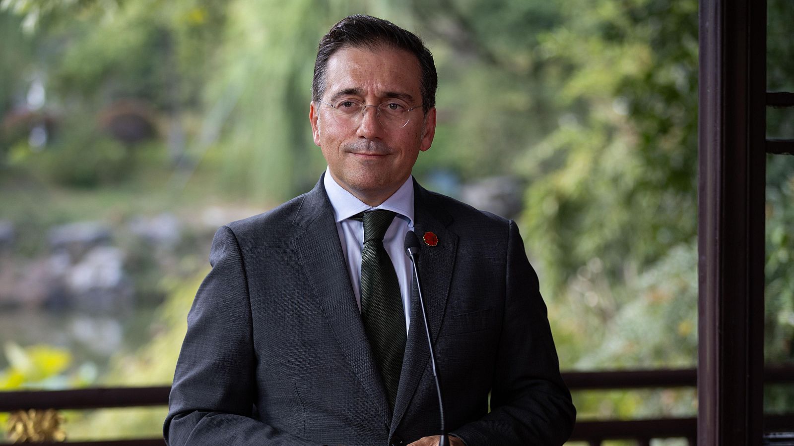 José Manuel Albares, el ministro de Exteriores, posa frente a un micrófono, vestido con traje oscuro, camisa blanca y corbata verde. Se encuentra en un jardín con vegetación y un estanque.
