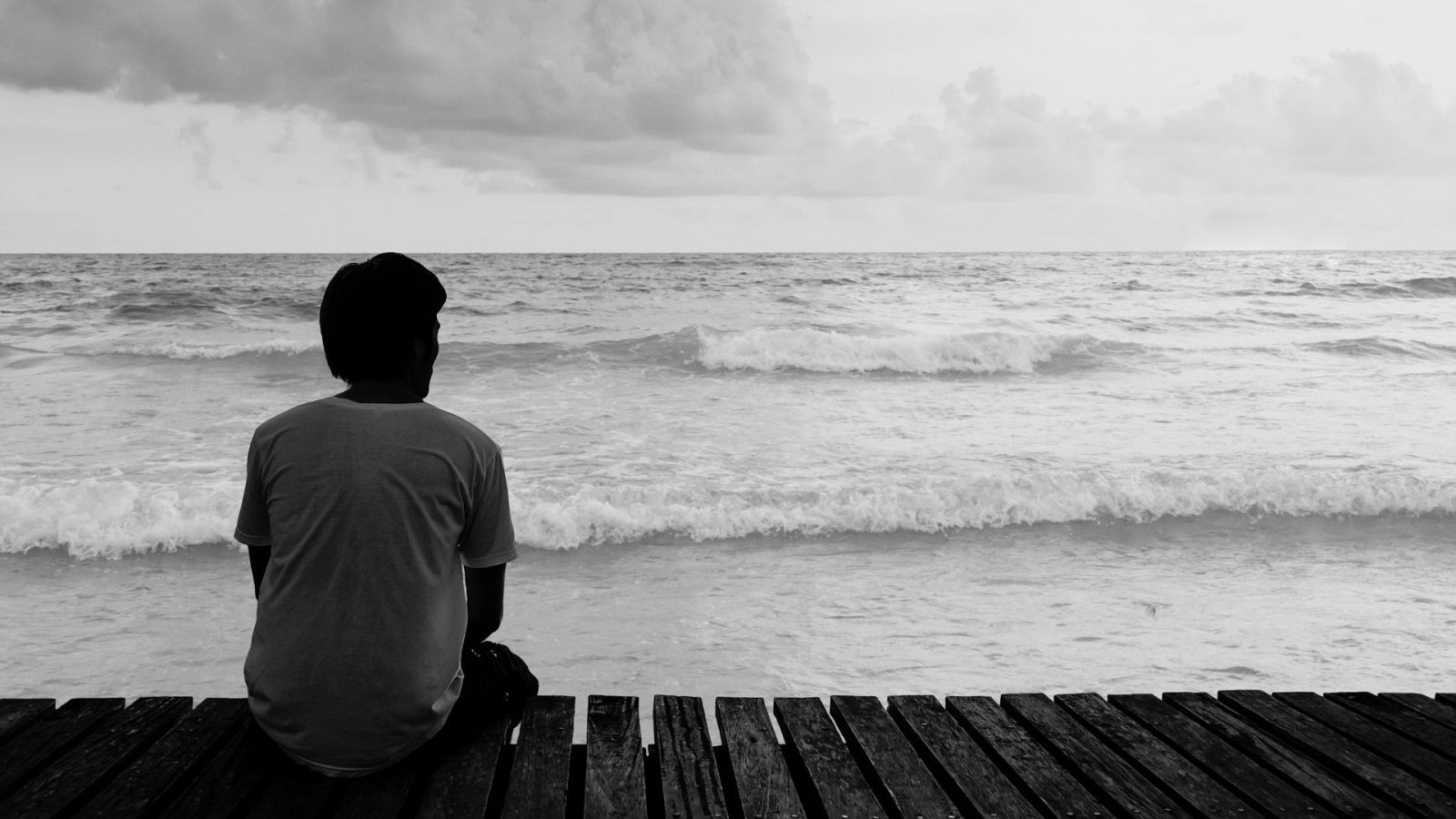 Persona sentada en un muelle, mirando un mar con olas bajo un cielo nublado.  Imagen en blanco y negro evoca soledad y reflexión.