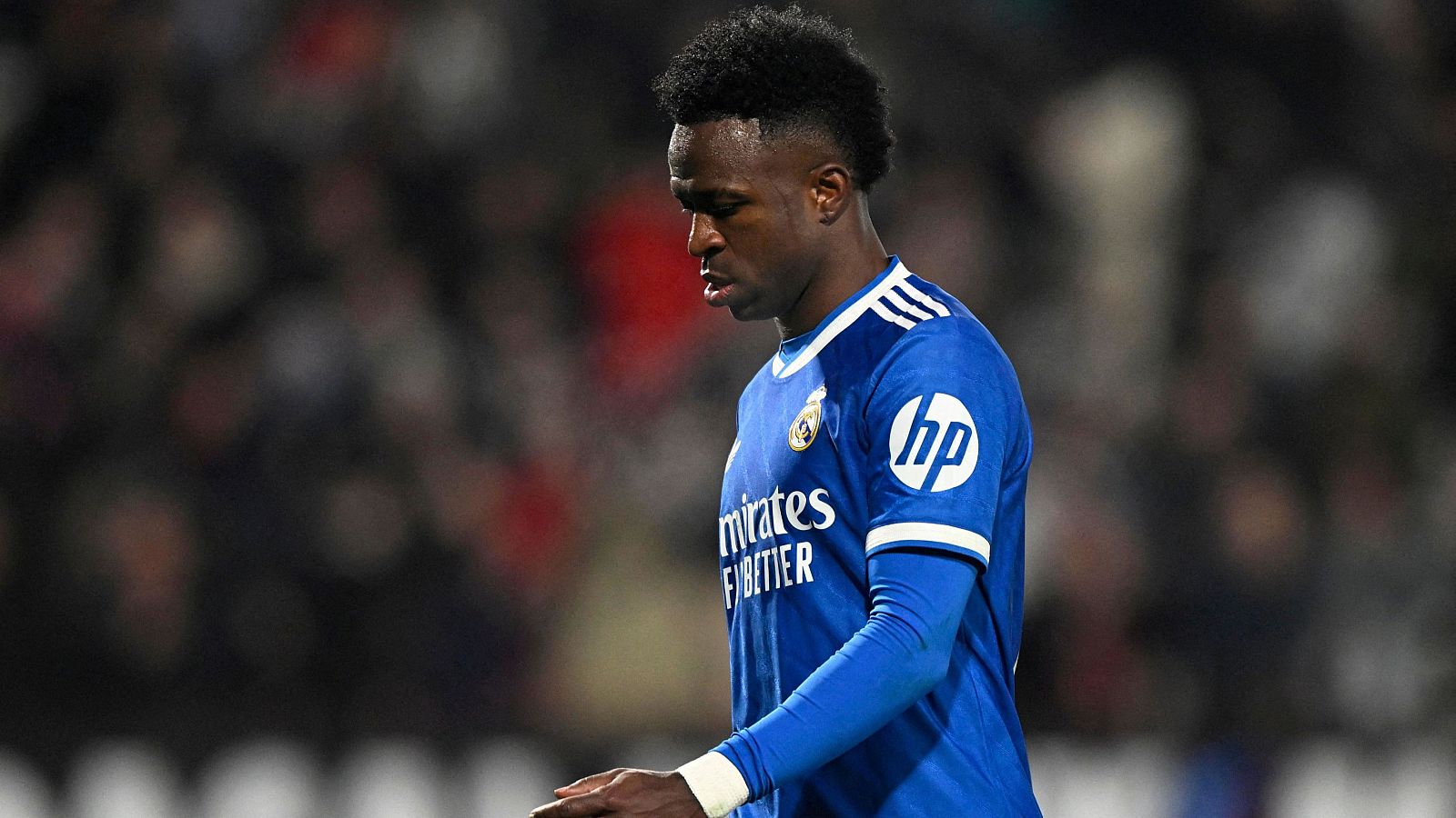 Vinicius, con camiseta azul del Real Madrid y mirada hacia abajo, posa en un estadio. Se distinguen los logos de los patrocinadores en su camiseta.