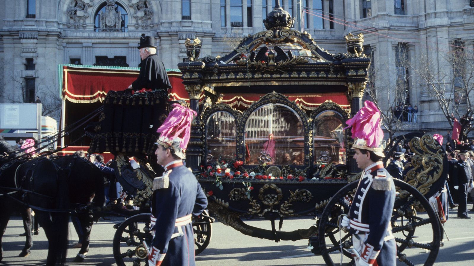 Una carroza fúnebre tirada por caballos, con un féretro en su interior, se desplaza frente a un edificio de estilo clásico. La escena incluye guardias uniformados y un hombre con sombrero de copa.