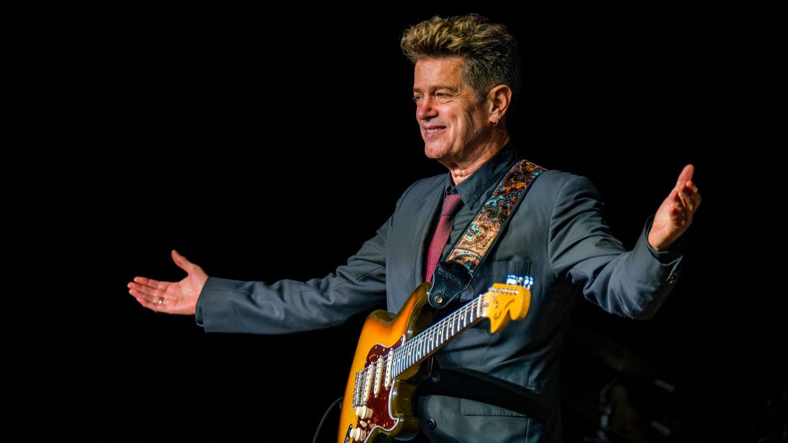 Santiago Auserón, con una guitarra eléctrica, sonríe a la cámara, vistiendo una chaqueta gris oscura, camisa oscura y corbata roja, con el fondo negro.