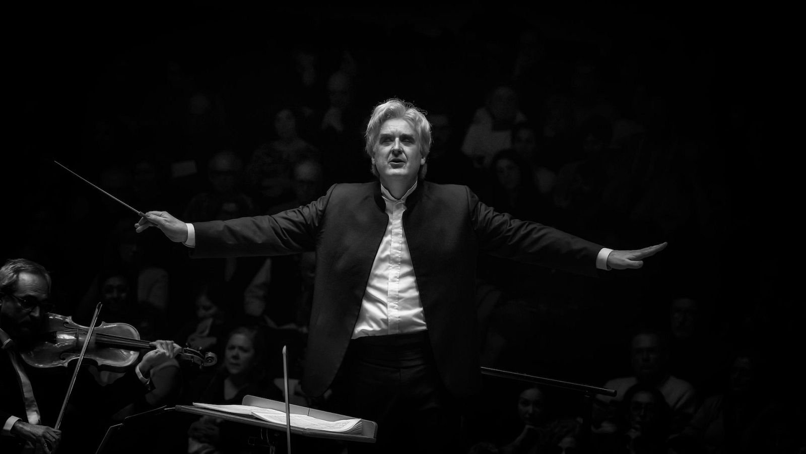 Thomas Dausgaard dirige a la orquesta, con los brazos extendidos y una batuta, en una imagen en blanco y negro. Los músicos tocan sus instrumentos, mientras el público observa el concierto.