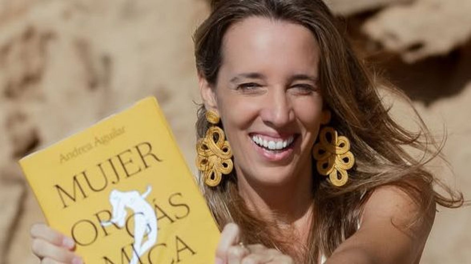 Una mujer sonriente, con pendientes dorados y cabello castaño claro, sostiene un libro con una portada amarilla y un título prominente, posiblemente en un entorno de color beige.