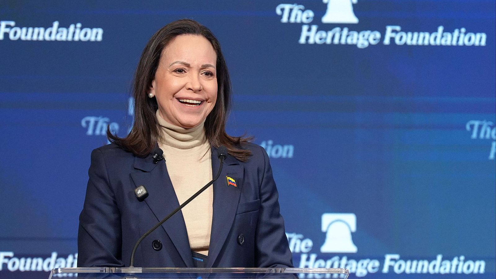 María Corina Machado, con un traje azul marino y un pin de la bandera venezolana, sonríe en una rueda de prensa. El fondo es azul con el logo de una fundación.