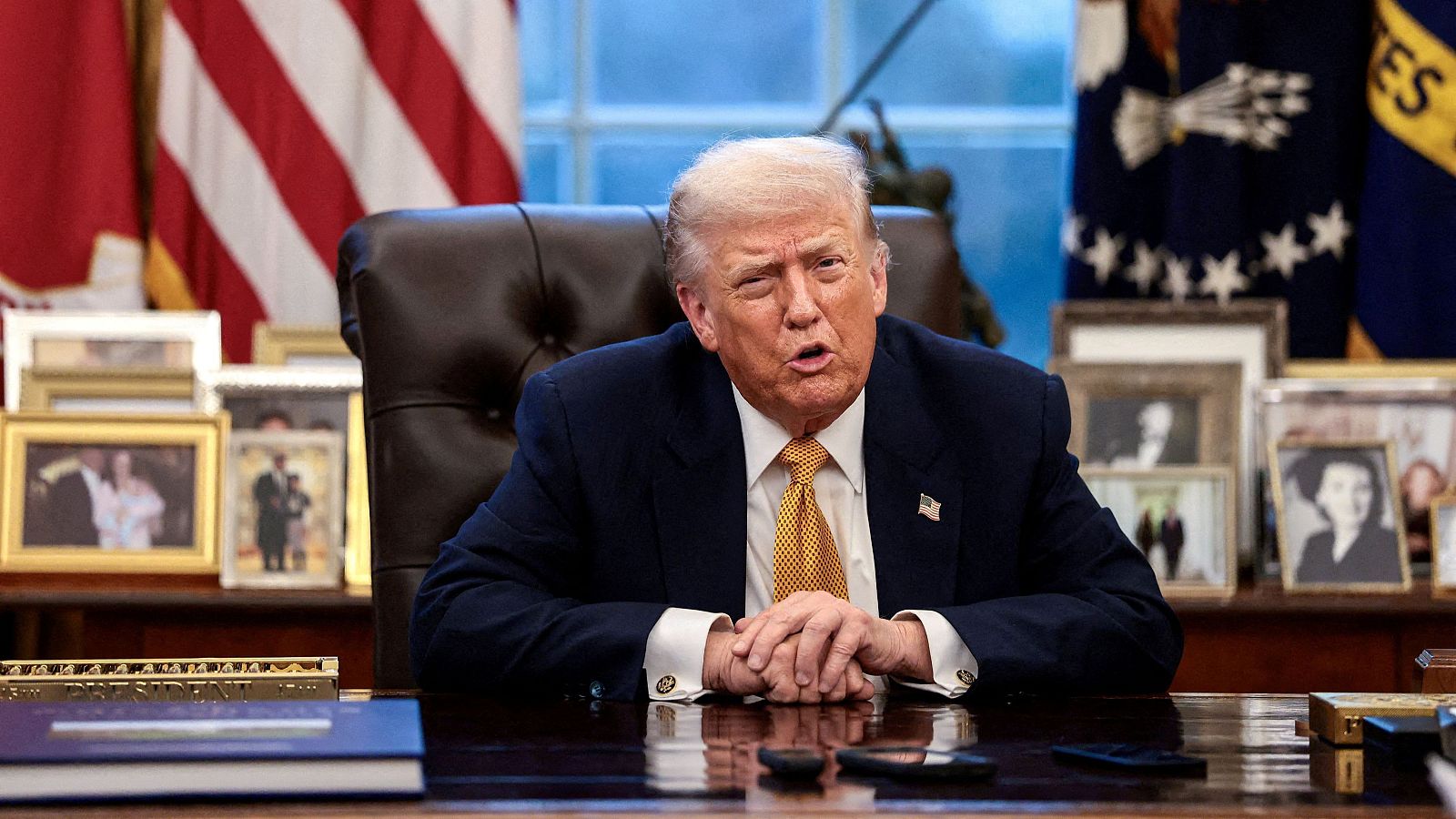 Donald Trump, vestido con traje oscuro y corbata, se encuentra sentado en un escritorio con las manos entrelazadas, con banderas y marcos de fotos detrás de él.