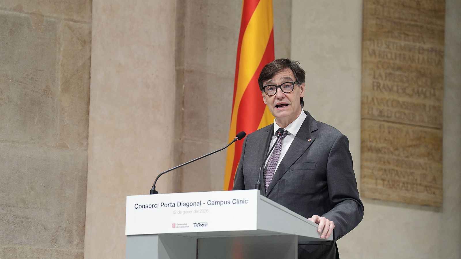 Salvador Illa, con traje y corbata, se encuentra frente a un podio con inscripciones en catalán y la fecha "12 de gener del 2024". A su lado, se observa la bandera catalana.