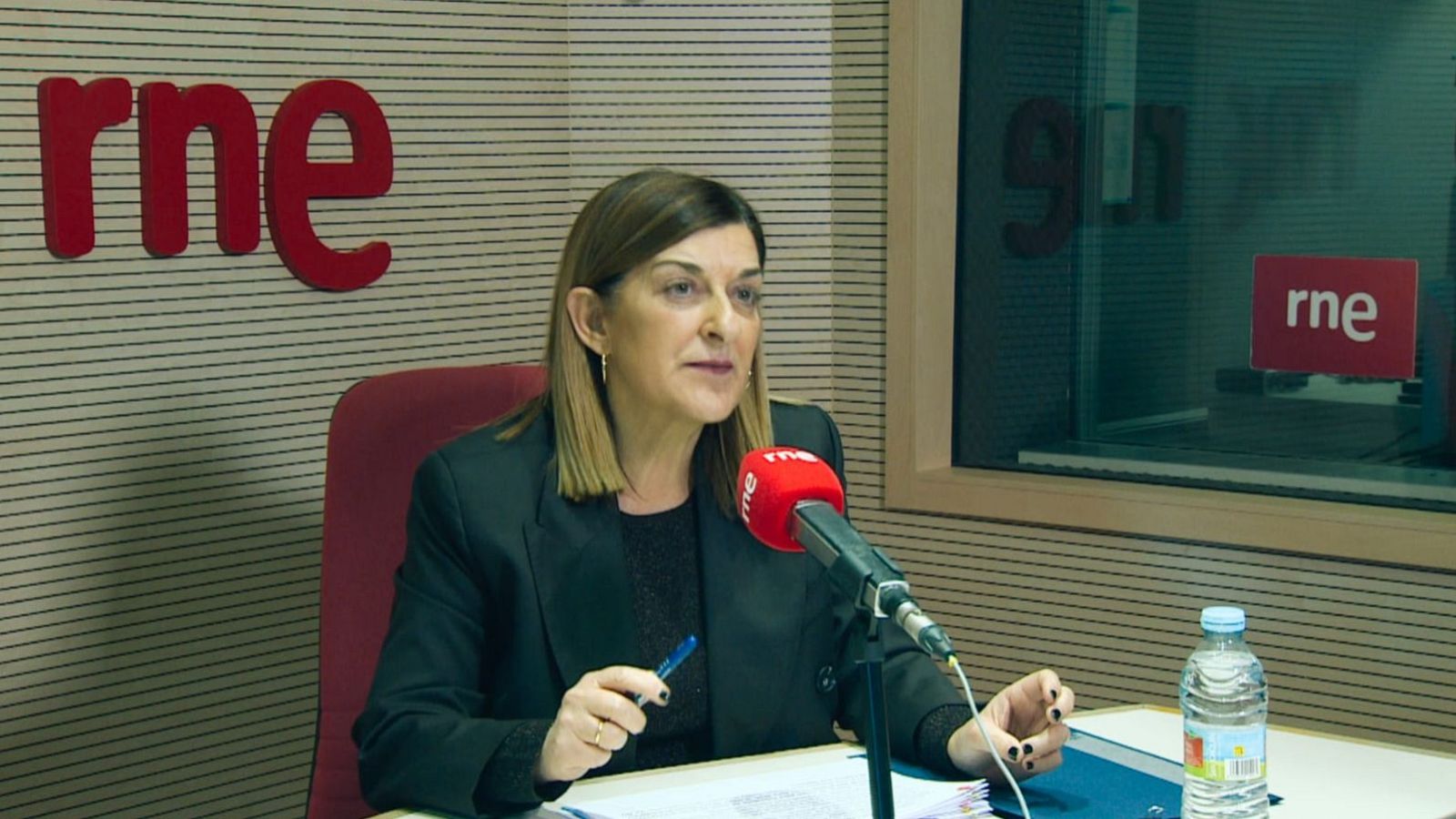 En un estudio de radio, una mujer con chaqueta negra y cabello castaño está sentada frente a un micrófono con el logo de RNE. Hay papeles, una botella de agua y una carpeta azul sobre la mesa.