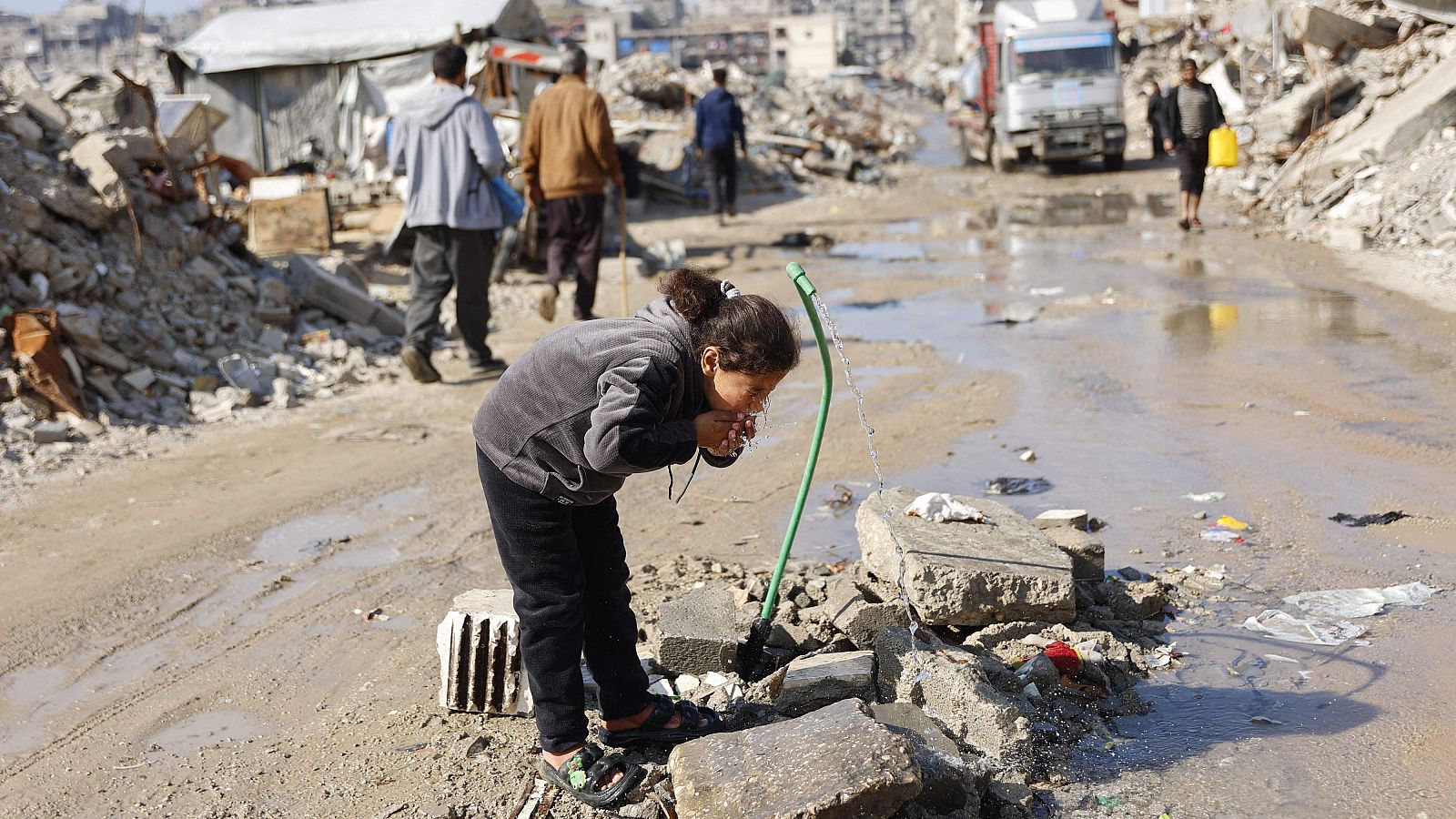 Una niña bebe agua de una manguera en medio de escombros. La escena muestra una niña con chándal gris y sandalias, en un entorno de desolación con restos de construcciones y agua estancada.