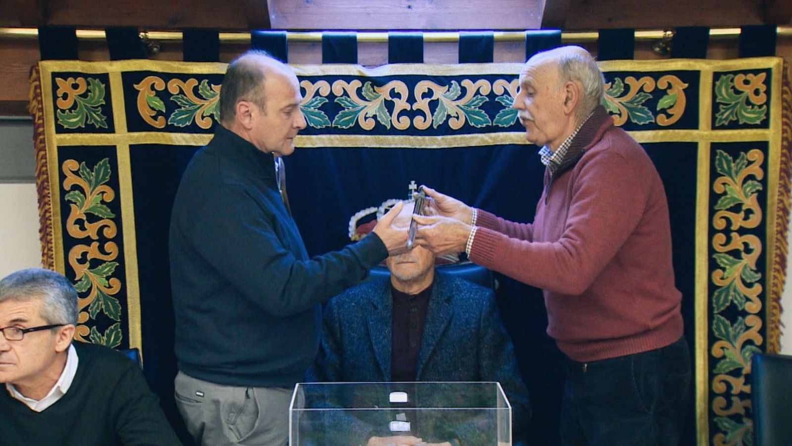 Un hombre con chaqueta oscura recibe un bastón de mando de otro con jersey rojo, en un acto oficial. Se encuentran frente a una mesa y un atril, con una tela decorativa oscura de fondo.