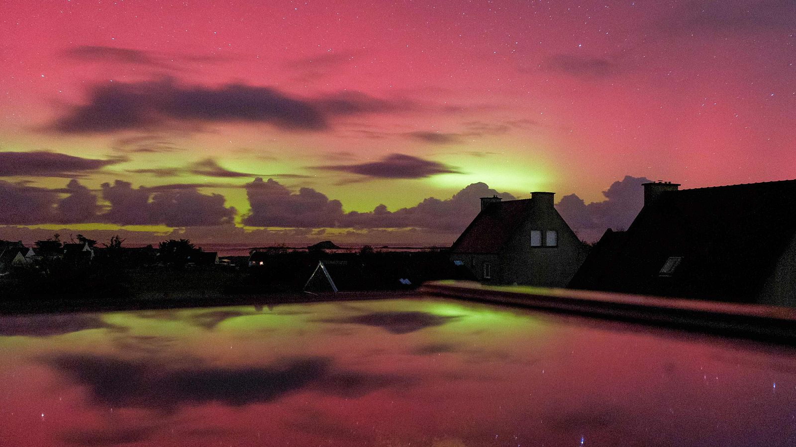 El cielo nocturno sobre Ploudalmezeau, Francia, se ilumina con una aurora boreal que despliega una banda de luz verdosa y amarillenta. Las siluetas oscuras de las casas se reflejan en una superficie lisa, creando un efecto visual simétrico y duplicado.