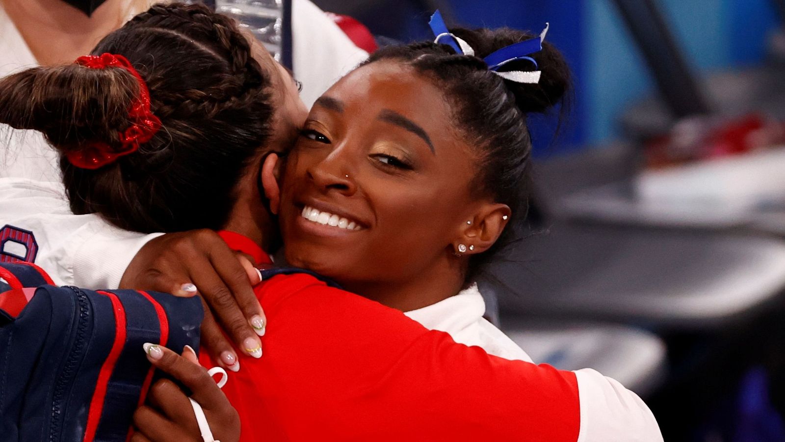 Simone Biles, con un moño y un uniforme deportivo, abraza a otra persona en una escena que parece ser de un evento deportivo. La otra persona también viste un uniforme deportivo.