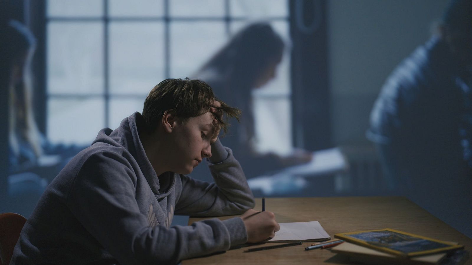 Un estudiante con una sudadera gris, aparentemente aburrido, se apoya en una mano con un lápiz, frente a un cuaderno abierto sobre una mesa de madera. El fondo muestra siluetas borrosas, sugiriendo un entorno de estudio.