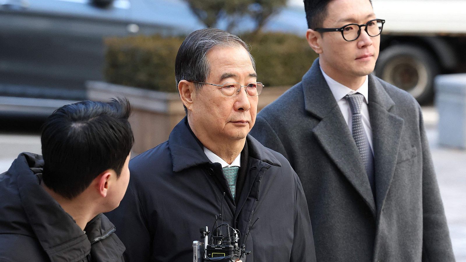 Fotografía de Han Duck-soo, un hombre mayor con gafas y abrigo oscuro, junto a otro hombre más joven, también con gafas y abrigo. Ambos llevan corbata y un micrófono.
