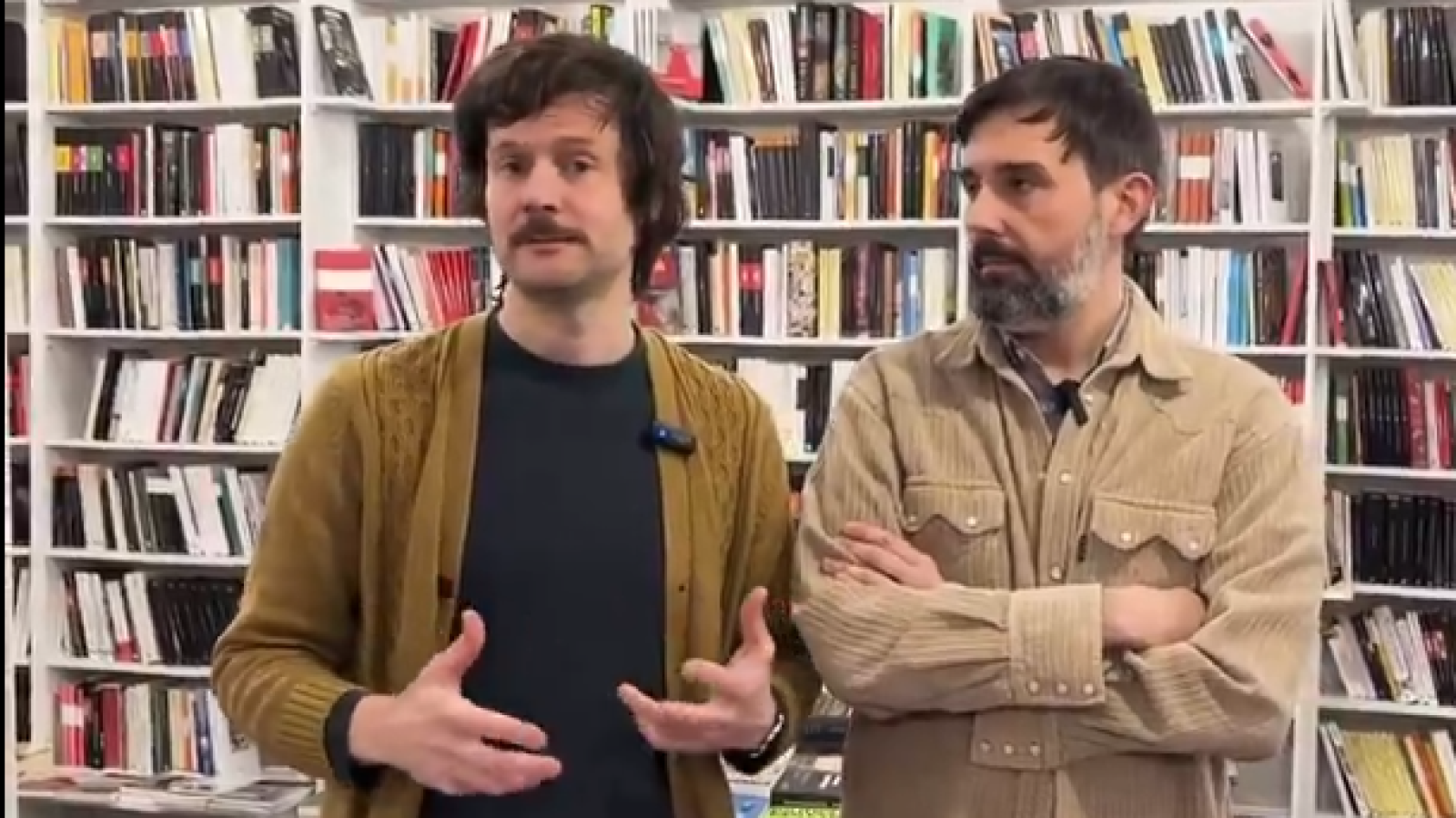 En una librería, dos hombres conversan frente a una estantería llena de libros. Uno lleva una chaqueta mostaza y un micrófono, mientras que el otro, con barba, tiene los brazos cruzados.