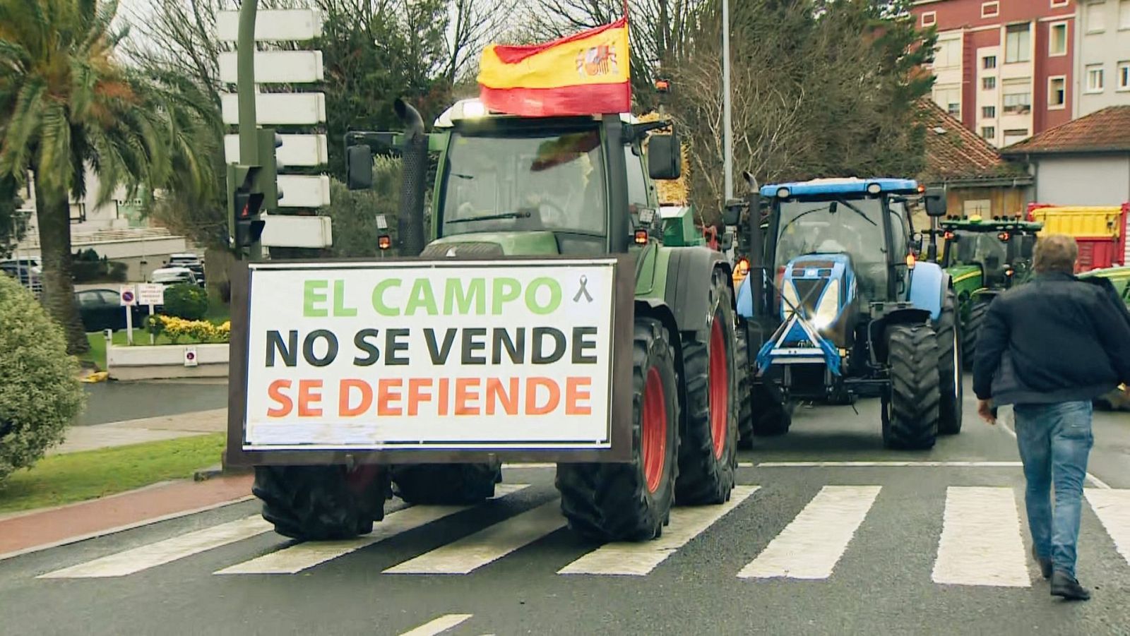 Una marcha de tractores, encabezada por uno que lleva una pancarta con un mensaje, recorre una calle de la ciudad. Se observa una bandera española ondeando sobre uno de los vehículos.