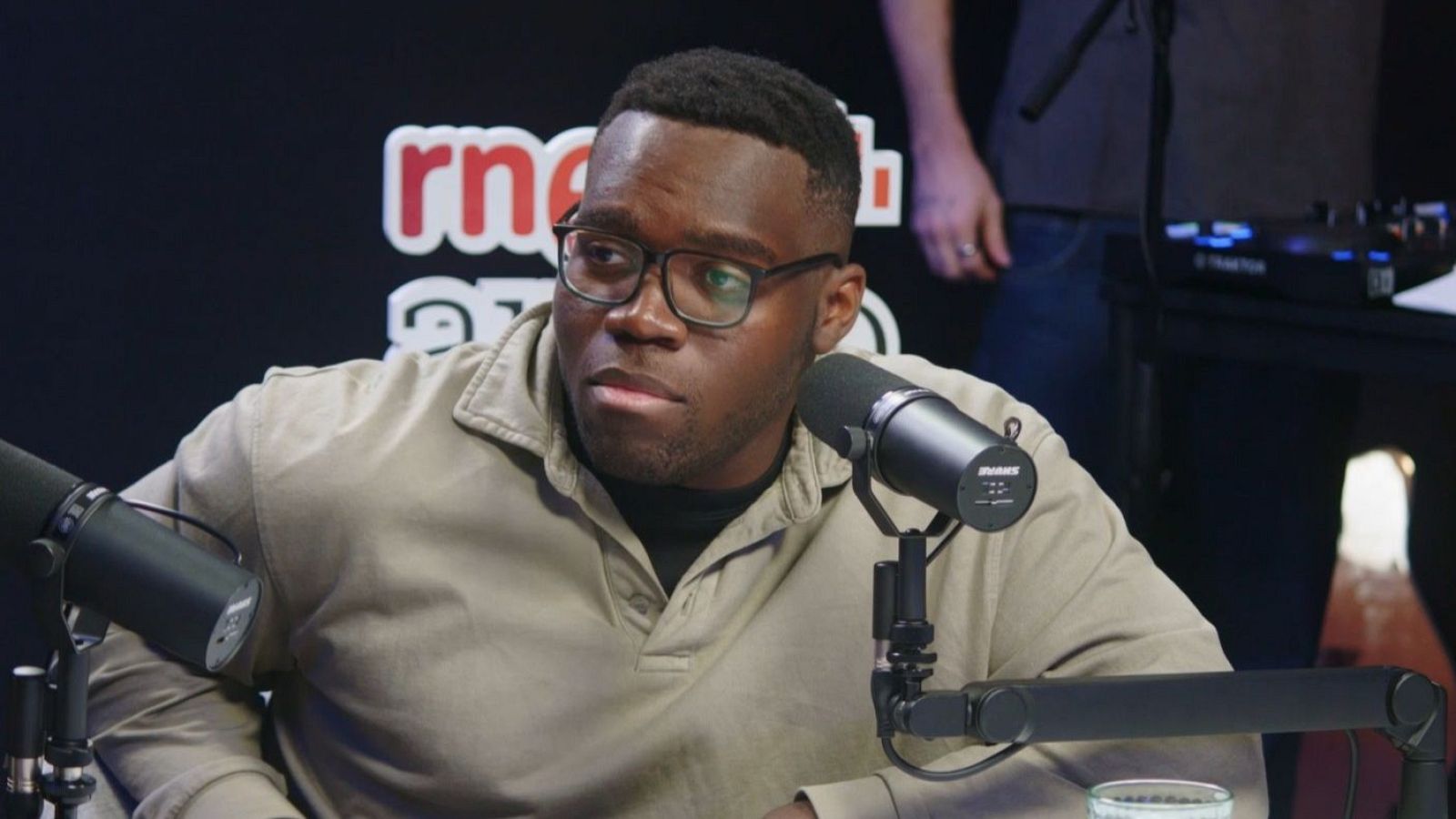 Un hombre con gafas y camisa beige se encuentra frente a dos micrófonos, posiblemente en un estudio de radio. Se aprecia el logo de una emisora con las letras "rn" y el nombre "ARKEBI Radio".