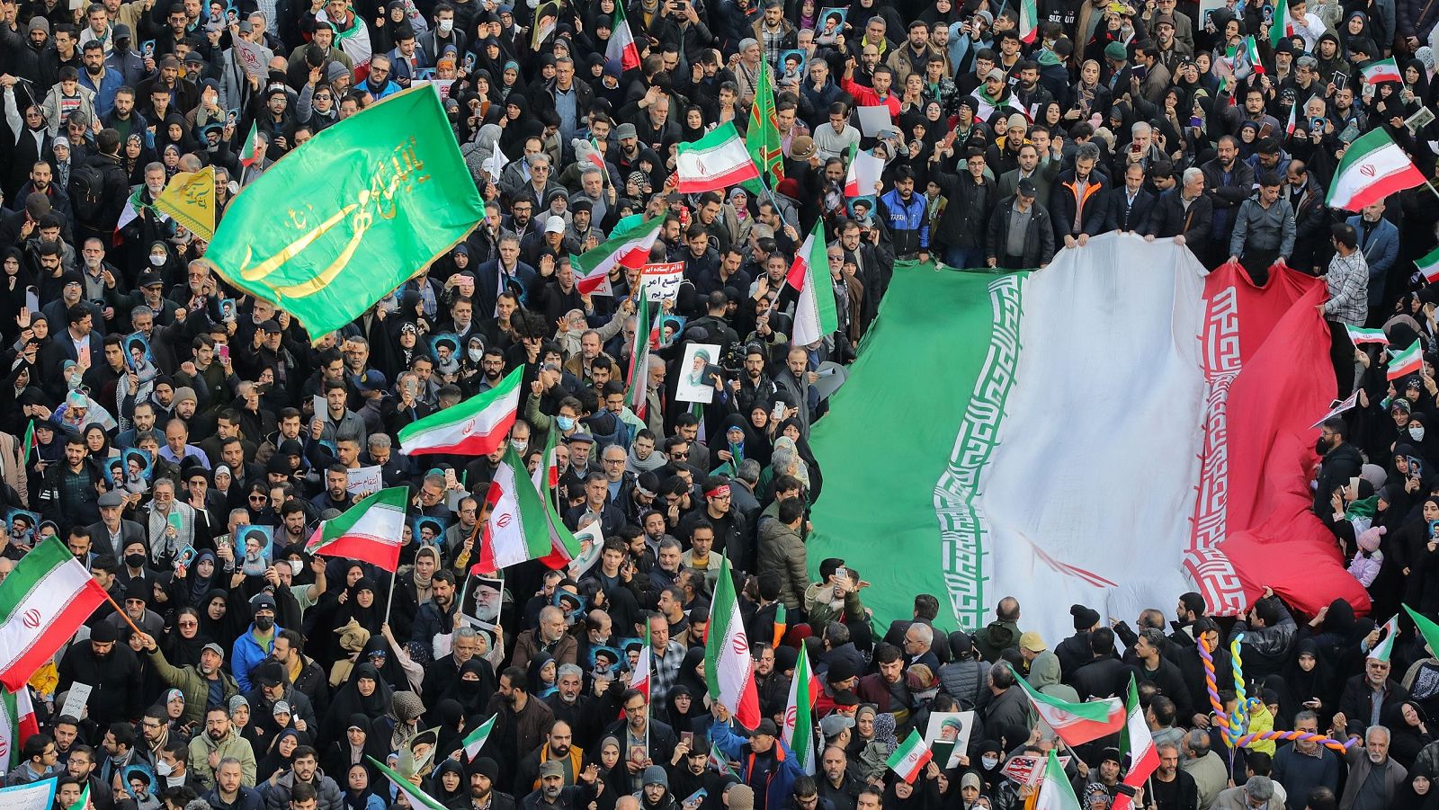 En una plaza de Teherán, una multitud de hombres y mujeres de diversas edades se manifiesta, ondeando banderas y sosteniendo pancartas. La atmósfera general sugiere una manifestación de apoyo.