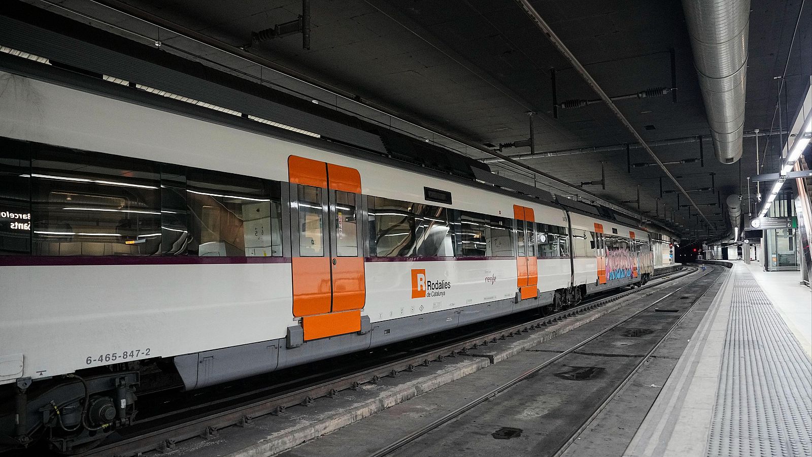 Un tren de cercanías blanco con detalles naranjas de Rodalies, con el logo visible y el número de serie 6-465-847-2, está estacionado en una estación subterránea con andenes grises y techo oscuro.