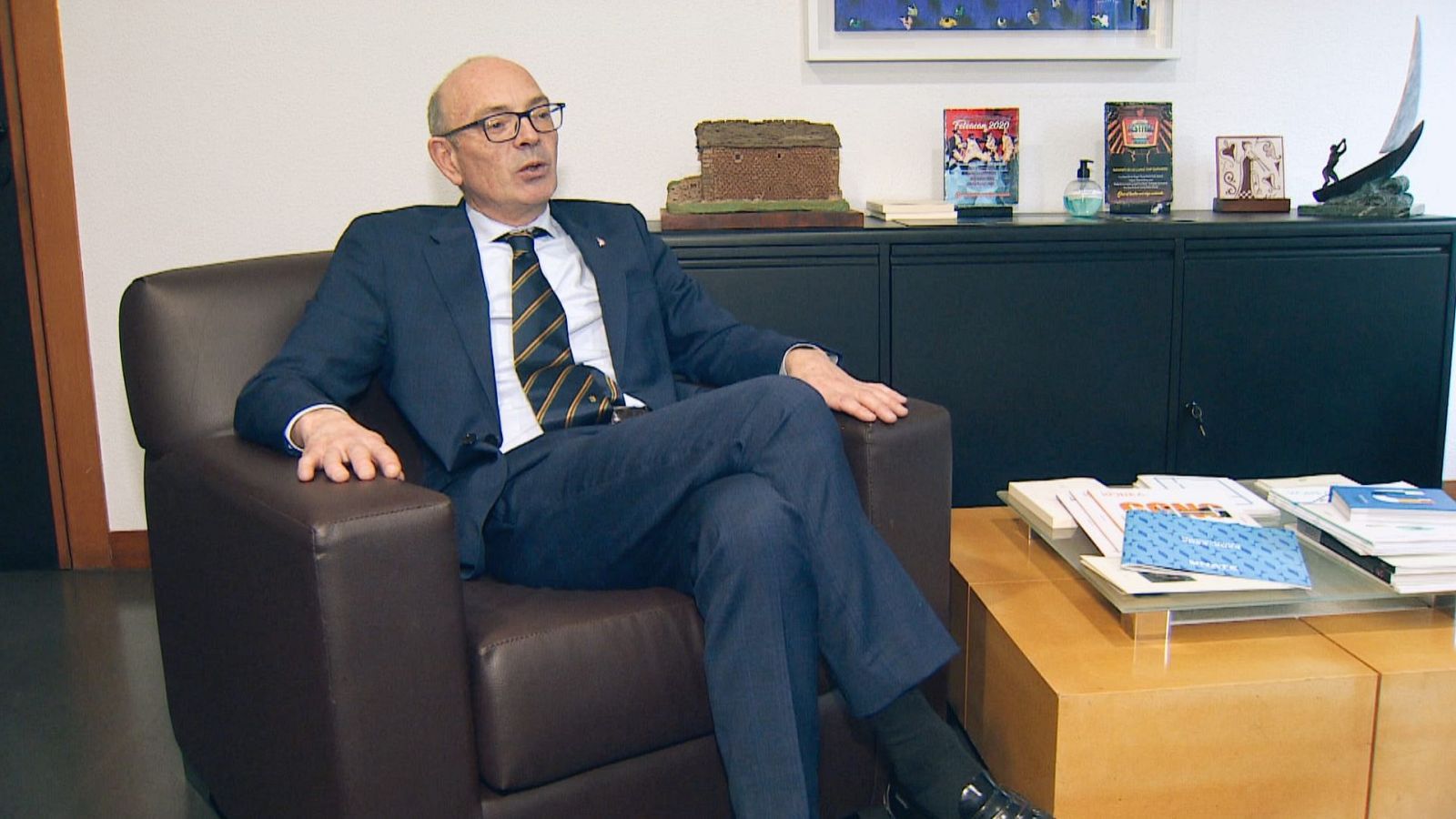 Ángel Pelayo, el nuevo rector, se encuentra sentado en un sillón de cuero en su despacho, con un traje oscuro y corbata. En el fondo, se aprecia un mueble con objetos y libros.