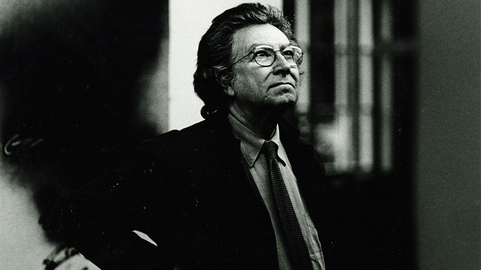 Retrato en blanco y negro de un hombre de mediana edad con gafas y corbata a rayas, mirando hacia arriba y a la derecha, en un interior con una ventana.