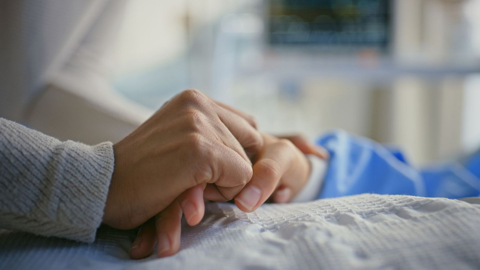 En una cama de hospital, dos manos se entrelazan. Una mano con manga de suéter gris sujeta otra mano cubierta por una sábana blanca, con un monitor médico borroso al fondo, evocando un ambiente de cuidado y apoyo.