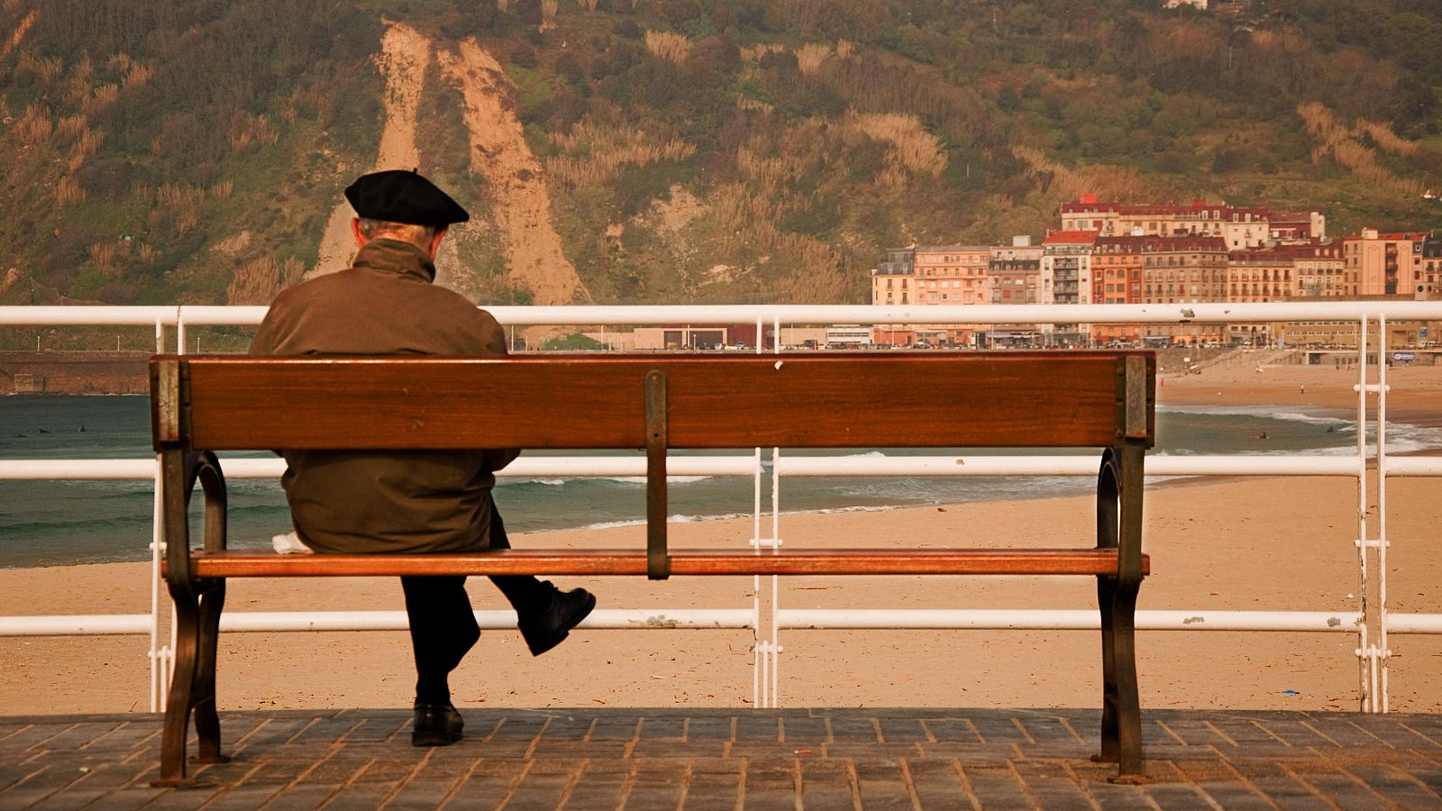 Un anciano con 'txapela' permanece sentado en un banco frente a una playa en San Sebastián