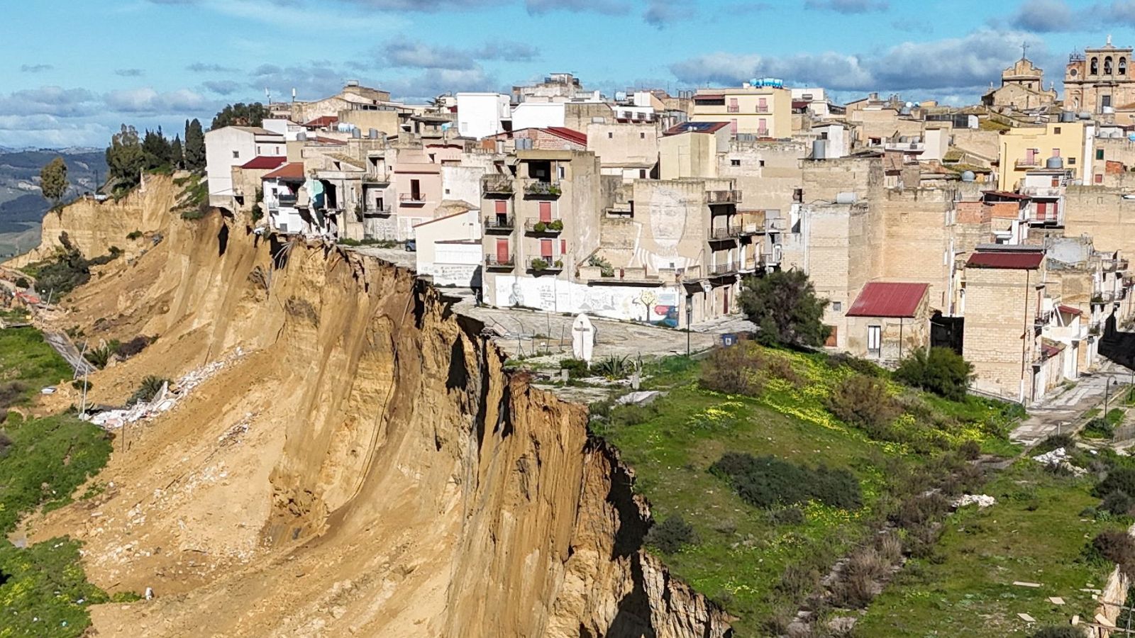 Un pueblo siciliano, con casas de piedra y balcones, se asoma al borde de un acantilado tras un deslizamiento de tierra. El terreno inestable revela desprendimientos y roca expuesta, con una iglesia visible en la parte superior.