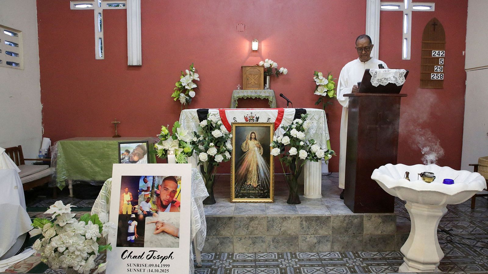 Un sacerdote, vestido con una túnica blanca, lee detrás de un atril en una iglesia. Frente a él, un altar adornado con flores y una bandera, con un cartel que muestra fotos y fechas. A la derecha, una pila bautismal con incienso.